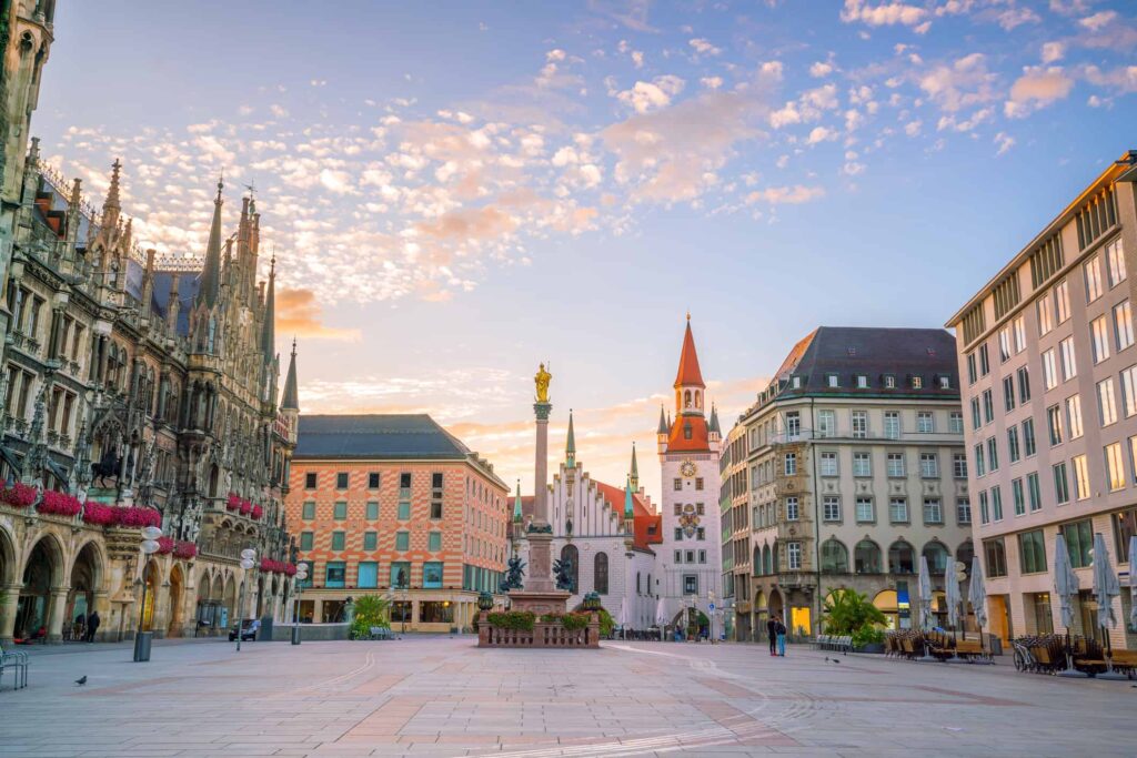 Image shows downtown Munich at sunrise. The image is of the Marienplatz flanked by stores and the old townhall.  