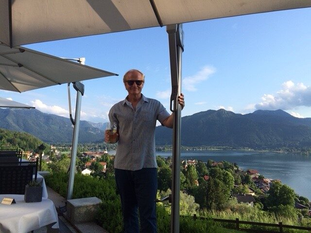 The image shows me standing in my little "quiet corner of the world." I am standing on the balcony of the Das Tegernsee hotel holding a glass of  "Tegernsee Hell." Behind me are the beautiful Bavarian mountains and lake Tegernsee.   